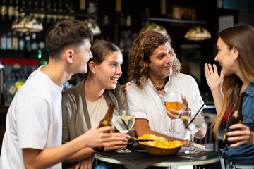 Group of happy carefree people spending time in pub, drinking beer with snacks and chatting in friendly way