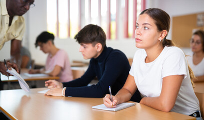 Young woman attending classes in college, sitting at desk and making notes. African-american man teacher answering young man's question.