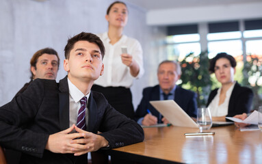 Portrait of young office worker sitting at table with colleagues, listening with interest and...