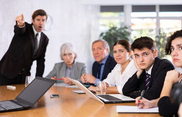 Frustrated young guy sitting with group of coworkers at table in director office on blurred...