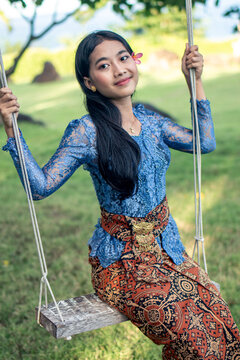 Balinese girl in traditional kebaya and batik sarong sitting on a swing in a tropical garden. 