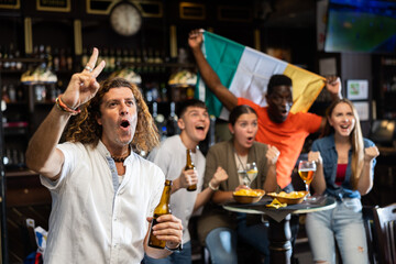 Happy adult football fan drinking beer and watching game on TV in sports bar proud of favorite team...