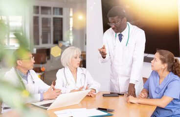 Middle-aged doctor talking to his colleagues sitting around table and listening to him