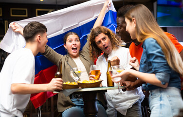 Happy multiracial sport fans holding up in a flag of Russia and watching football competitions in the bar
