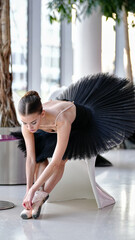 Fototapeta premium Focused ballerina in a black tutu leans forward to adjust her pointe shoes, sitting on a sleek white chair in a bright dance studio with natural light