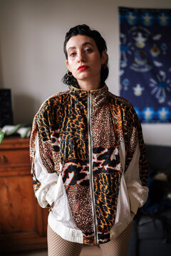Pensive alternative female with red lips wearing tights jacket in leopard print looking at camera and standing with hands in pockets at home