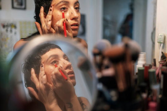 Concentrated informal female with nose piercing applying eyeliner on lower eyelid while looking in mirror in dressing room