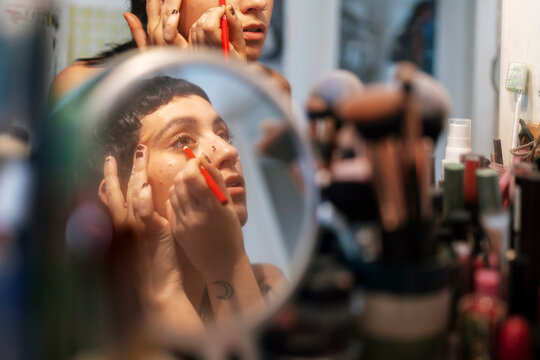 Crop focused informal female applying eyeliner on lower eyelid and looking in vanity table mirror in dressing room