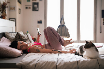 Side view of young tattooed female browsing smartphone and touching cat while resting on bed in light bedroom in morning at home