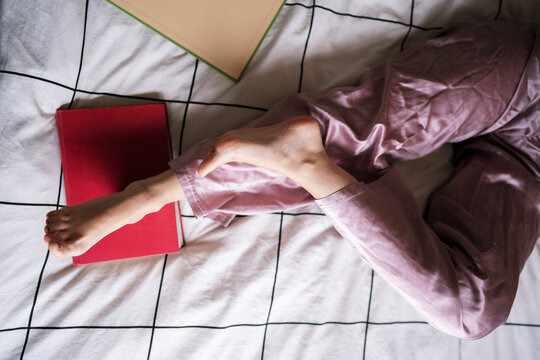 Top view of anonymous barefoot female in silk sleepwear lying on bed near books and relaxing at home