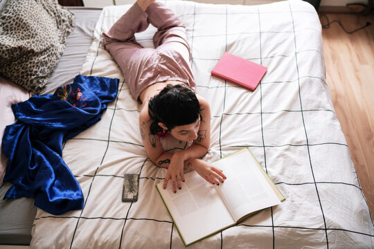 From above tattooed young female in sleepwear relaxing on bed and reading book in morning at home