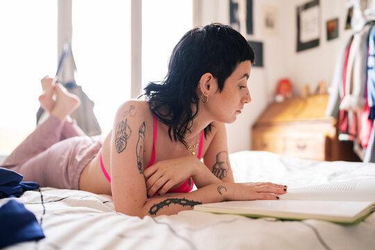 Informal young woman with tattoos adjusting bra and reading interesting book while chilling on bed in morning at home