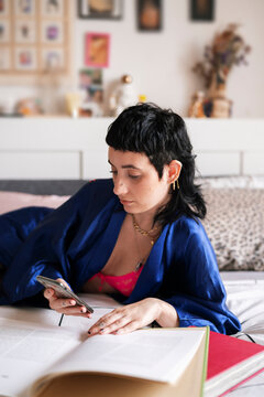 Informal young woman in silk nightwear using mobile phone and reading book while lying on bed in light bedroom