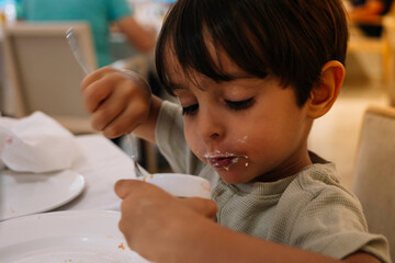 A young boy is eating a bowl of ice cream with a spoon. He is wearing a green shirt and has messy hair