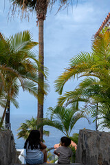 Two children are sitting on a stone staircase overlooking the ocean. The scene is peaceful and serene, with palm trees in the background and the sound of the waves in the distance