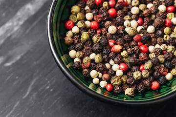 Mixed peppercorn spices in bowl. Colorful seasoning on black stone background. Red, white, green, and black pepper. Top view of spice mix. Aromatic cooking ingredients. Natural flavor concept.