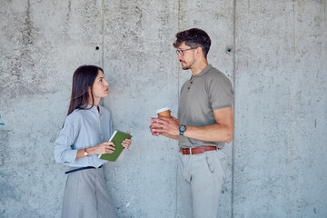 Two young entrepreneurs talking about new business ideas during coffee break, standing against concrete wall