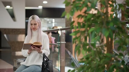 poised woman seated on bench flipping book pages with two hands, wearing cozy sweater, jeans and boots, black bag beside, mother and daughter entering moving walkway behind glass railing - Powered by Adobe