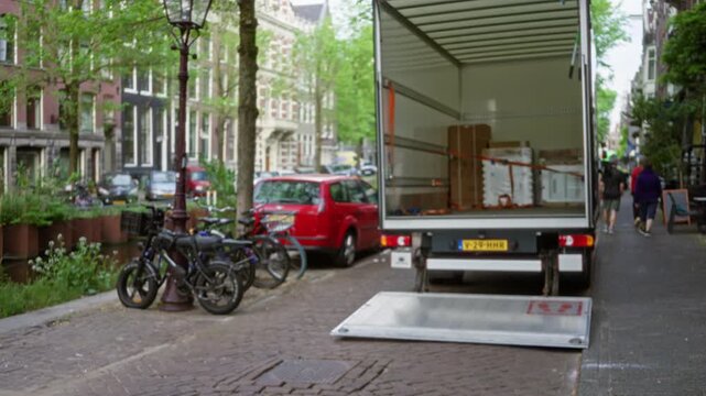 Truck parked on amsterdam street with open tailgate, bicycles, and walking people in blurred background, showcasing urban lifestyle and european architecture.