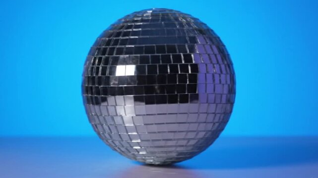 A silver disco ball rests on a gray surface set against a blue backdrop The balls surface is covered in small mirrored tiles