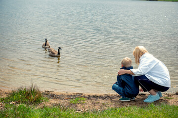 Young blonde grandmother hugs her grandson while sitting by a mountain lake. Peaceful moment, ducks...