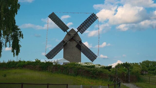 Old wooden windmill standing in a grassy rural landscape beneath a blue sky with scattered clouds, captured during a sunny day. The surroundings evoke a sense of tranquility and history.