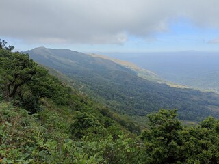 Lush mountain landscape showcases expansive greenery and distant sea views under a cloudy sky at midday