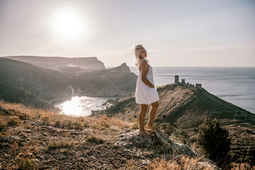 Naklejka premium A blonde woman stands on a hill overlooking the ocean. She is wearing a white dress and she is enjoying the view.