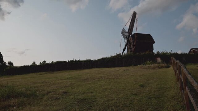 Camera Panning Along Traditional German Wooden Windmill in Picturesque Countryside Landscape Under Blue Sky. The surroundings evoke a sense of tranquility, history, and scenic beauty. 