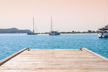 A wooden pier extends into a calm turquoise sea. Several sailboats are anchored in the distance....
