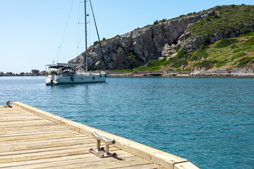 A wooden pier extends into a calm blue sea. A white sailboat is anchored nearby. Rocky cliffs and...