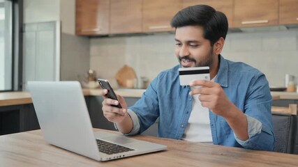 Smiling young man making an online payment with a credit card and smartphone while seated at a wooden table in a cozy kitchen, working on his laptop in a comfortable home setting
