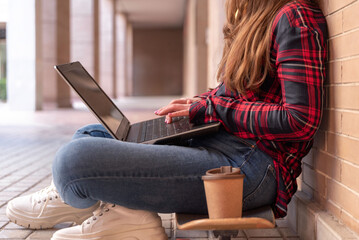 Close up of female student using laptop while sitting on skateboard on college campus with coffee