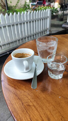 A cup of hot espresso served with a glass of melted sugar, and a large glass of ice water on a wooden table looks luxurious. Natural blur background, in outdoor cafe.