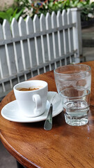 A cup of hot espresso served with a glass of melted sugar, and a large glass of ice water on a wooden table looks luxurious. Natural blur background, in outdoor cafe.