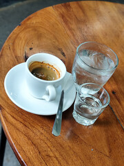 A cup of hot espresso served with a glass of liquid sugar, and a large glass of ice water on a wooden table, in an outdoor cafe.