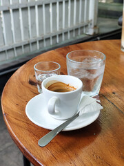 A cup of hot espresso served with a glass of liquid sugar, and a large glass of ice water on a wooden table, in an outdoor cafe.