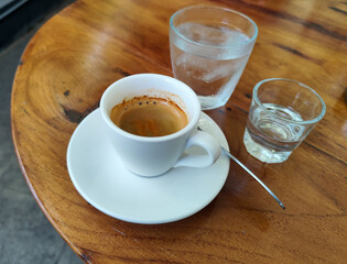 A cup of hot espresso served with a glass of liquid sugar, and a large glass of ice water on a wooden table, in an outdoor cafe.