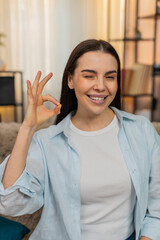 Young happy woman at home on sofa confidently showing OK gesture with hand smiling in positive...