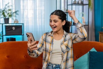 Indian young woman at home sitting on sofa with mobile phone joyfully screaming, celebrating...