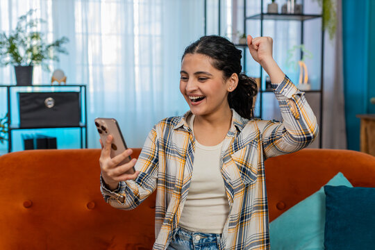 Indian young woman at home sitting on sofa with mobile phone joyfully screaming, celebrating lottery win or good news. Arabian girl emotionally expressing triumphant excitement, happiness and joy