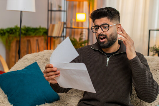 Indian man at home opens envelope, reads letter and looks upset by bad news, reacting with sadness. Arabian guy sits on sofa disappointed by rejection of job, credit, or failed exam results in letter.