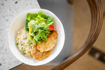 Homemade meal served in a white bowl with oatmeal, green peas, fresh salad leaves, and crispy fried patties on a speckled surface for a healthy food concept.