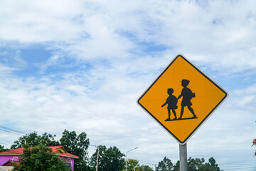 Bright yellow school crossing warning sign against a cloudy sky evokes safety. Students shown represent education and childhood, creating an image symbolic of vigilance and care.