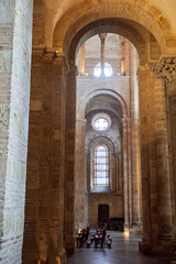 Stone arches and vaulted ceilings tower over wooden pews in a Romanesque church interior, lit by...