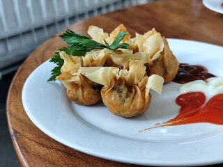 Crispy fried dumplings served with celery leaves, as well as chili, tomato and mayonnaise sauce arranged artistically on a white plate on a wooden table in an outdoor cafe. Natural blur background.