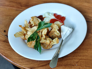 Crispy fried dumplings served with celery leaves, as well as chili, tomato and mayonnaise sauce arranged artistically on a white plate on a wooden table in an outdoor cafe. Natural blur background.
