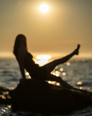 Silhouette of person sitting on rock near sea with leg raised during sunrise. Relaxation and summer vacation concept for design and poster