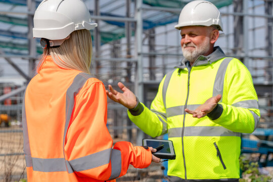 Two construction professionals, man and young woman, engaged in conversation at construction site, both wearing safety helmets and high-visibility jackets while reviewing plans on a tablet