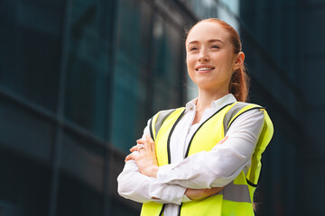 Young woman in safety vest smiles confidently in front of modern office building made of steel frame and dark glass